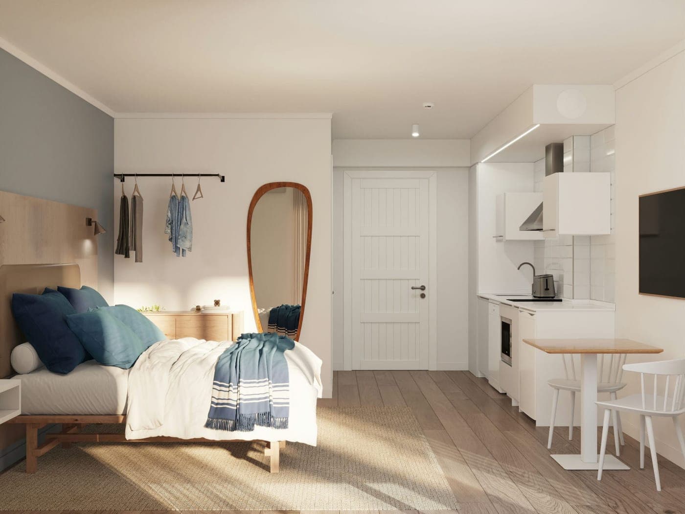 Interior de un estudio con cama doble, perchero, espejo ovalado y una cocina americana con una mesa pequeña
